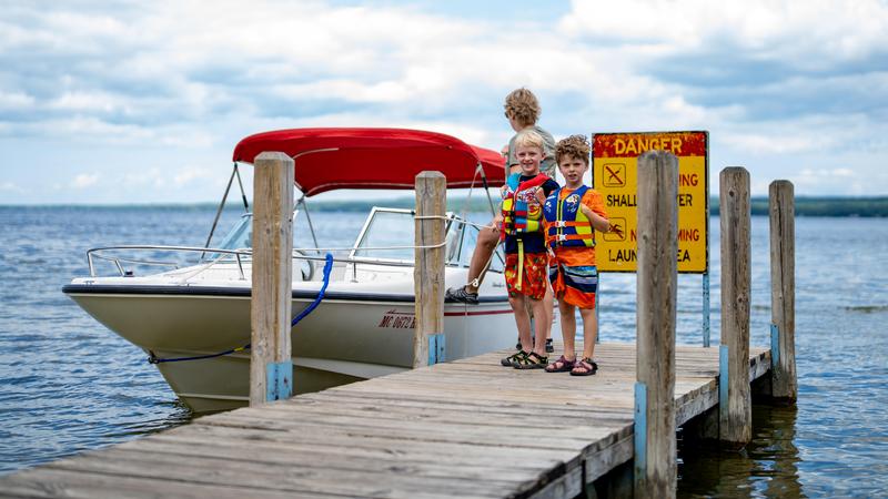 Two children with lifejackets on stand on a pier at a boat launch with a boat tied to the posts ready to launch