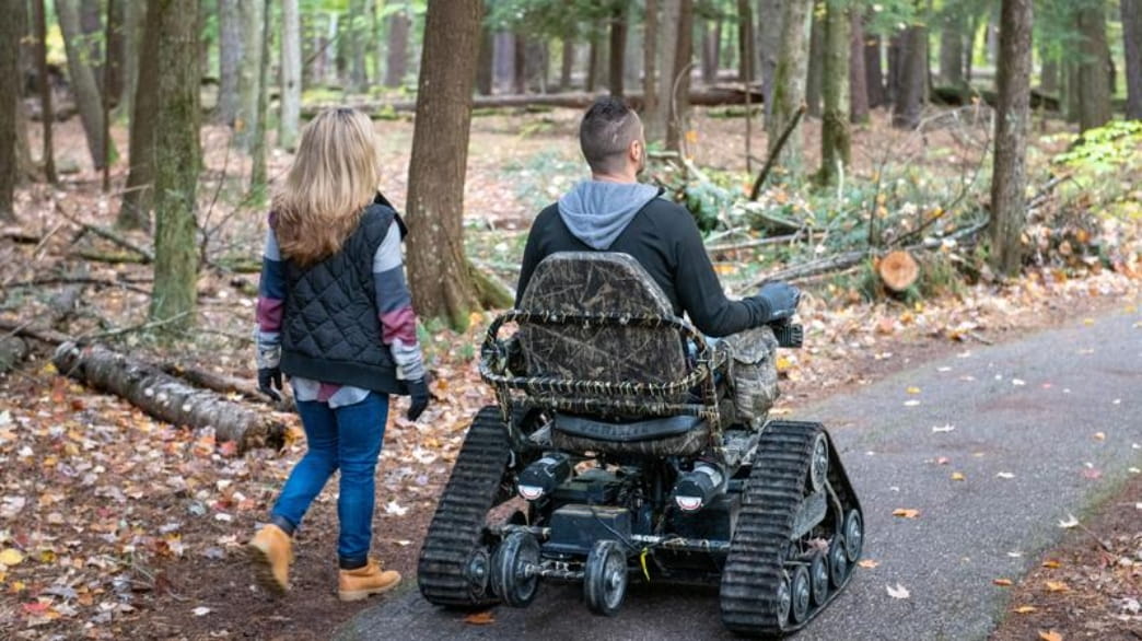 Two people travel on a path in the woods, one is using a track chair
