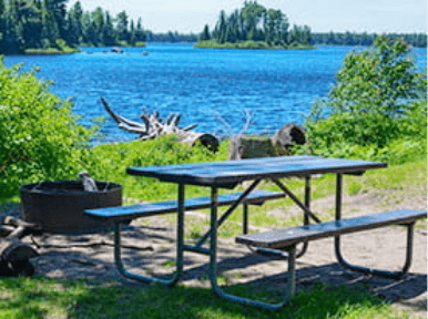 A picnic table sits by a lake with a firepit in the background