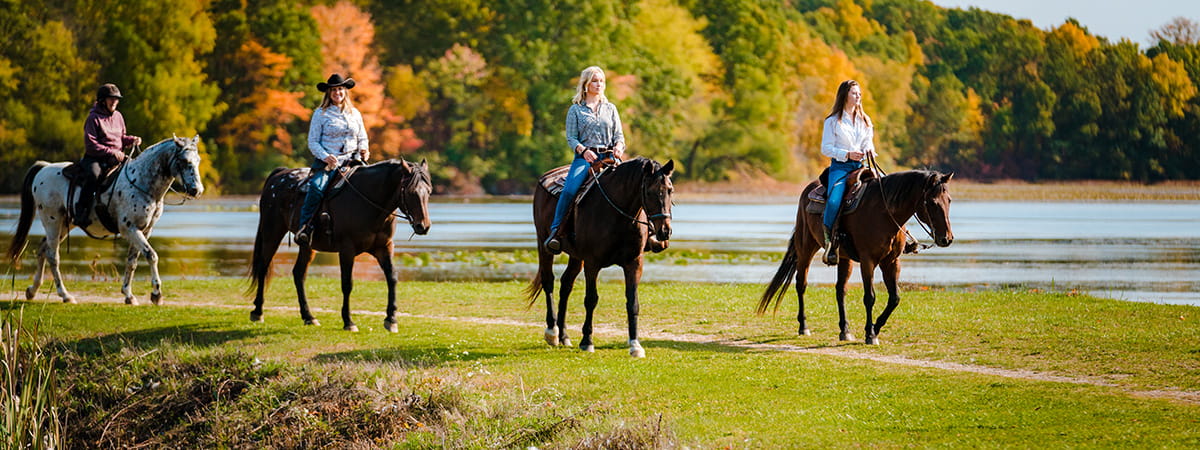 A group riding horses on a trail passing by a pond with fall colors on the trees