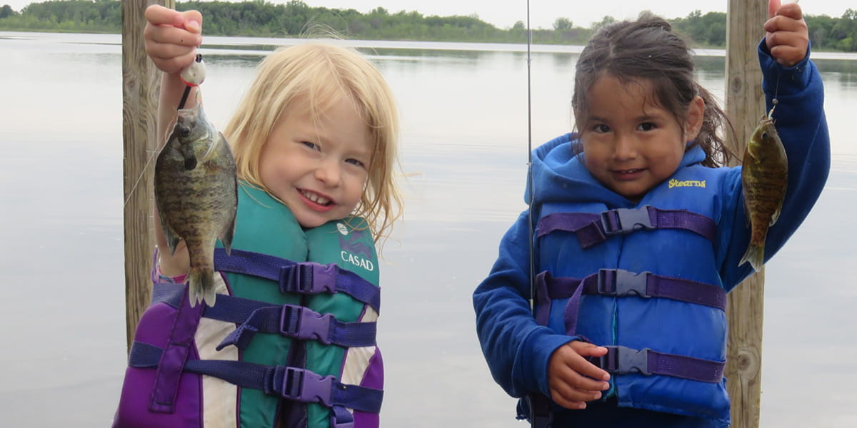Two girls standing on a dock with life jackets on holding up fish they caught