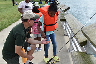 An educator shows a child how to fish. They are standing on a fishing pier and the girl is wearing a life jacket.