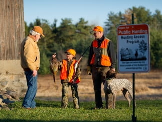 A young girl about 10 years old wearing hunter orange and cradling a shotgun in the crook of her arm holds up a recently harvested pheasant to show a landowner.