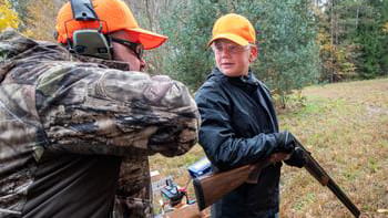 A youth hunter receiving instruction from an adult while he holds an unloaded shotgun in a forest clearing. 