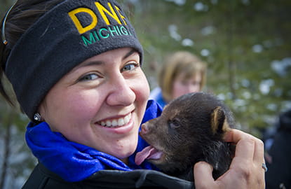 A DNR worker holds a bear cub in her hands while smiling as the bear yawns.