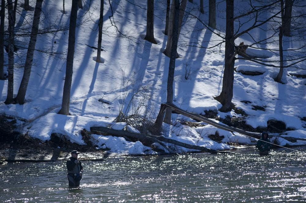 An angler stands in a river fishing with snow on the ground in the background