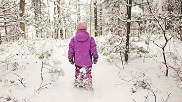 A girl dressed in bright pink snowgear stands on a snowy trail in her snowshoes
