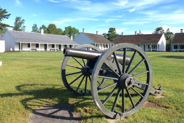 A large, wheeled cannon sits on a large grassy area. White one-story wooden buildings are in the far background.