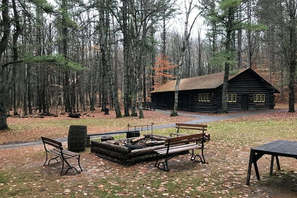 Benches surround a squared-off firepit area in the woods. In the background, a long log building is nestled between trees. 