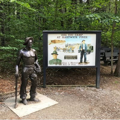 A bronze statue of a young man in a hat and with a shovel sits next to a large sign about the CCC.