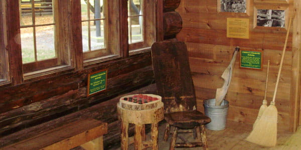 In a corner of a log building, a wooden chair sits next to a handmade checker board. 