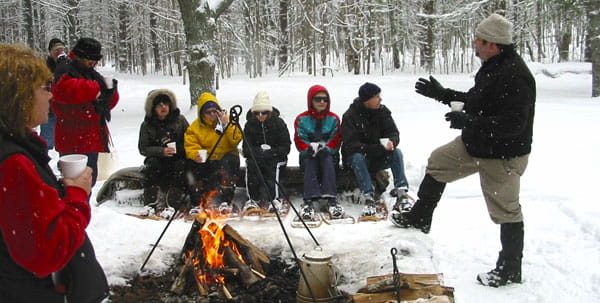 6 people in winter cloths and snowshoes sit on a snowy log in front of a campfire. A standing man talks to them. 