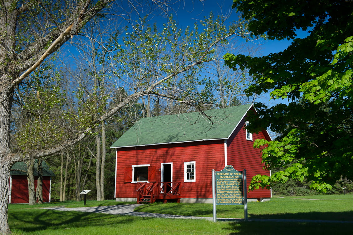 A green and gold historical marker sits in front of a red wooden work building. 