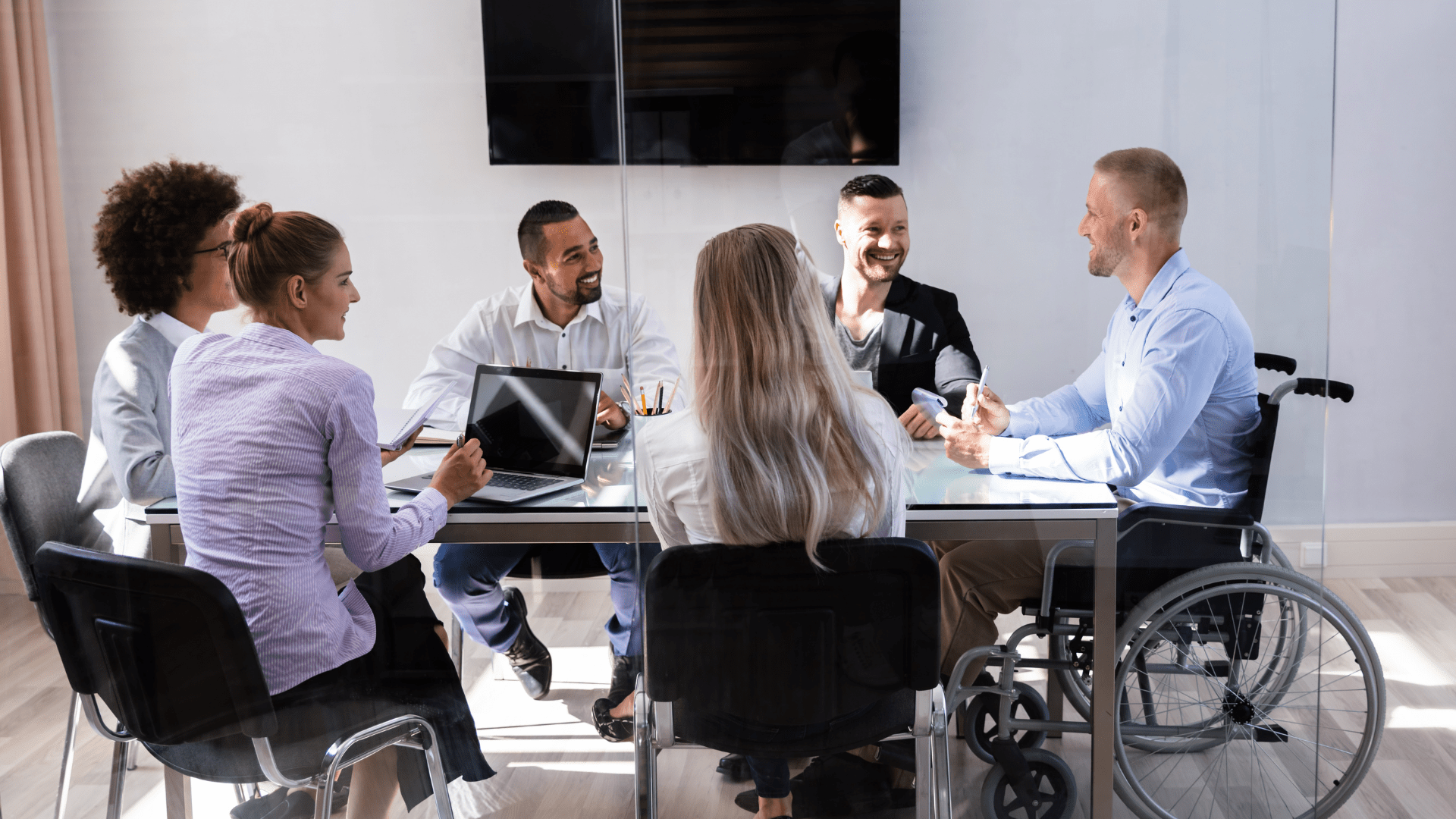 Image of a people meeting, sitting at a conference room table. Several people are sitting in chairs while one member is seated in a wheelchair. 