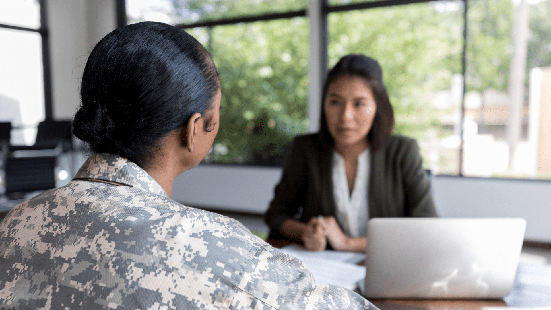 Image of a woman dressed in military attire speaking with another woman at a desk. 