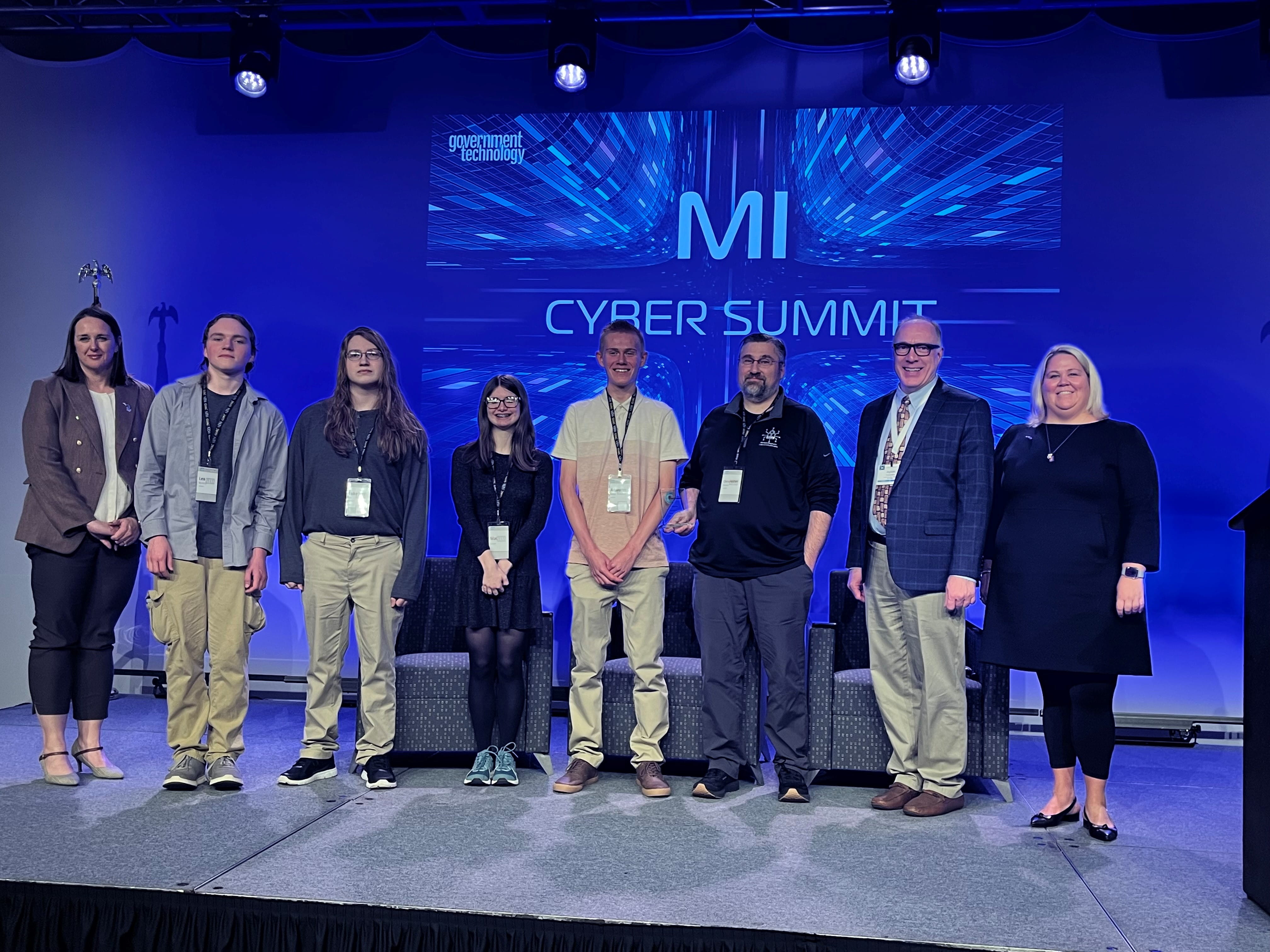 Five students with three representatives from DTMB on a stage with "MI Cyber Summit" in the background.