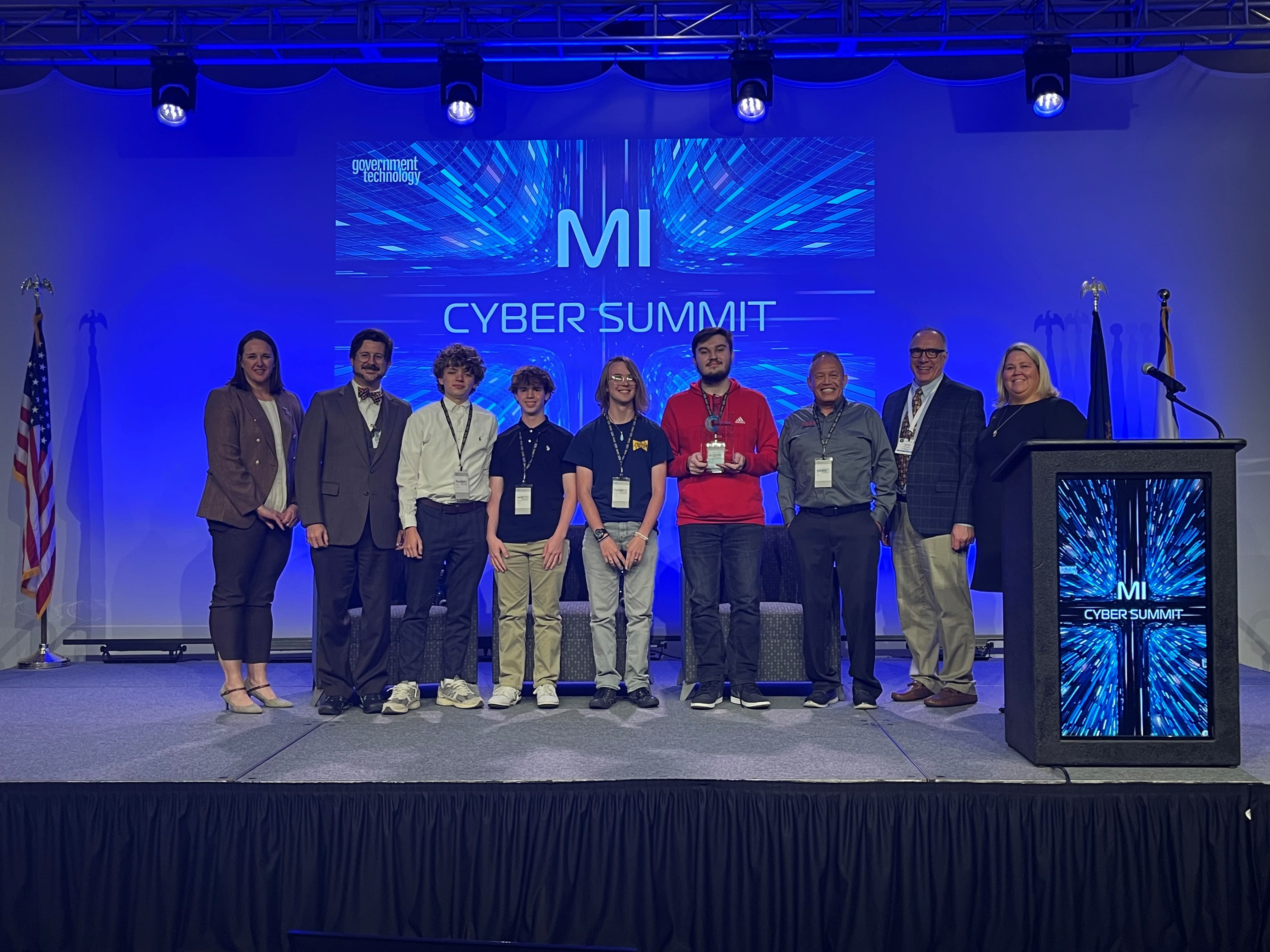 Students, coaches and DTMB representatives on a stage with "MI Cyber Summit" in the background.