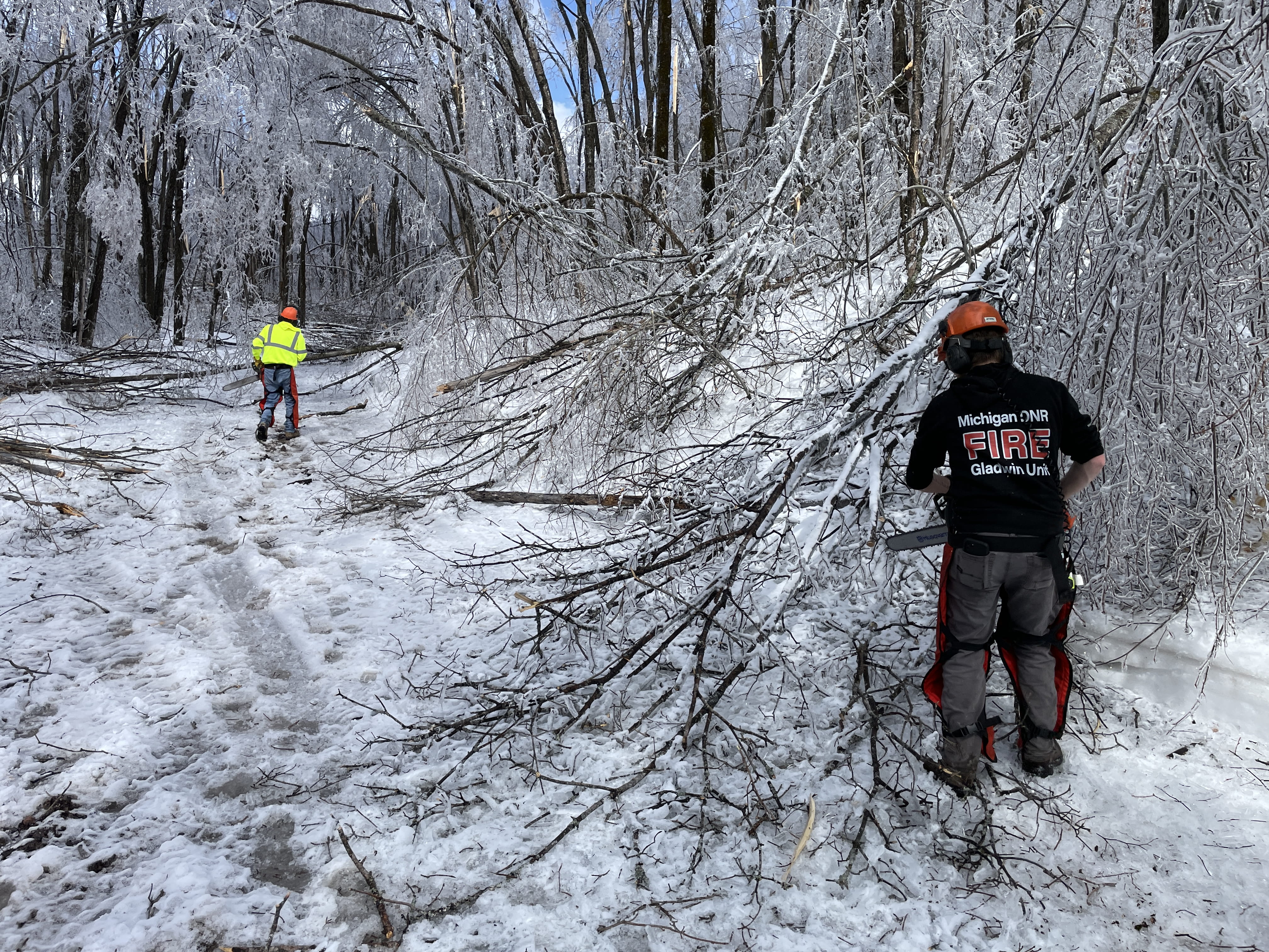 Two first responders using chainsaws to clear ice covered trees bocking a road.