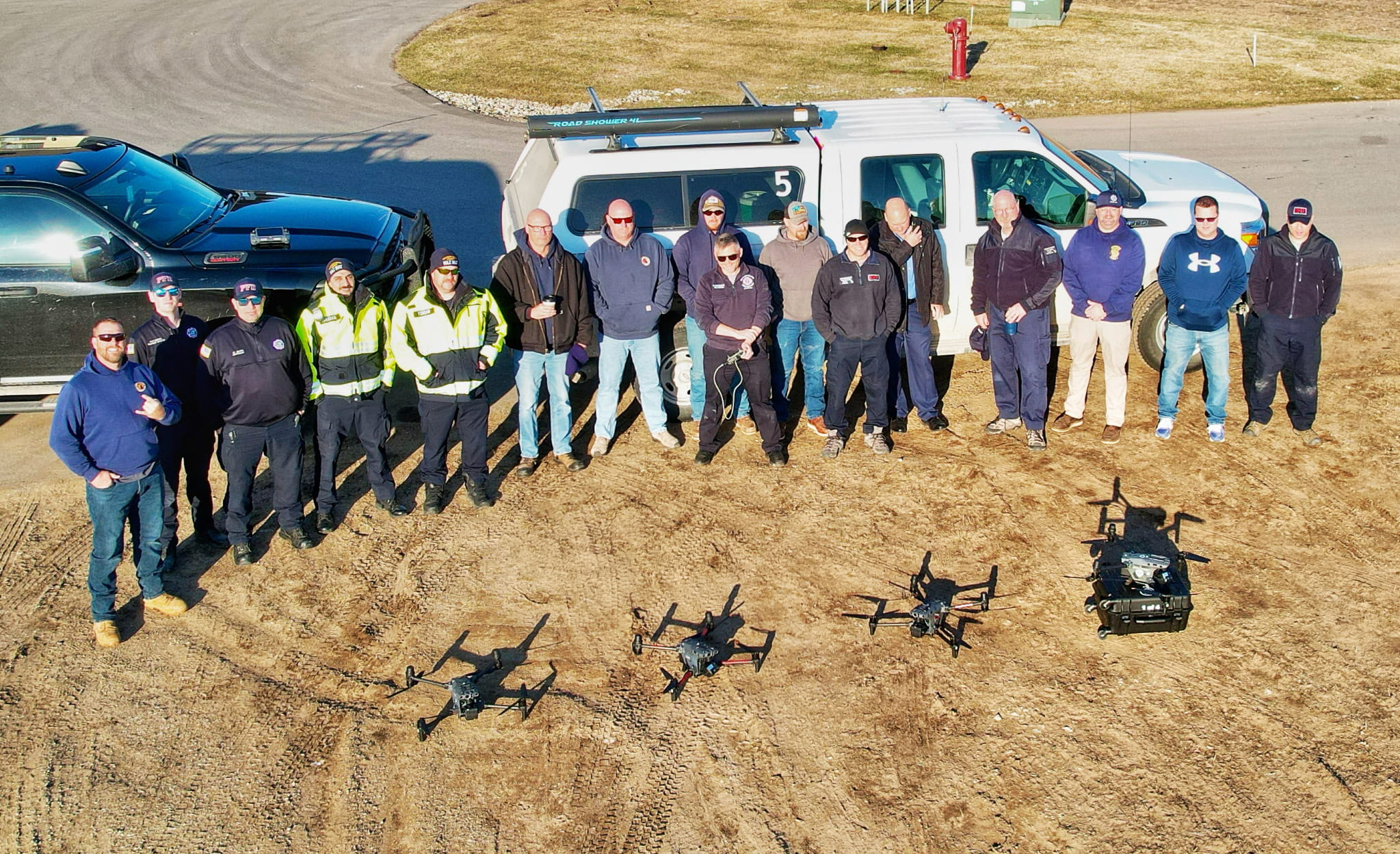 Sixteen individuals posing in front of four drones.
