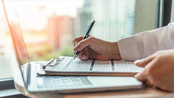 Photo of hand holding a pen filling out a form sitting on a laptop against a window glowing with orange sunlight.