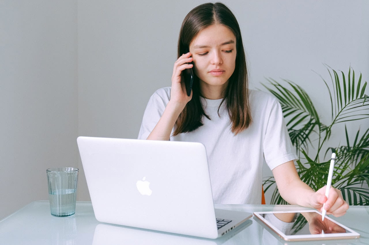Woman talking on phone while taking notes on a tablet. There is an open laptop on the table in front of her.