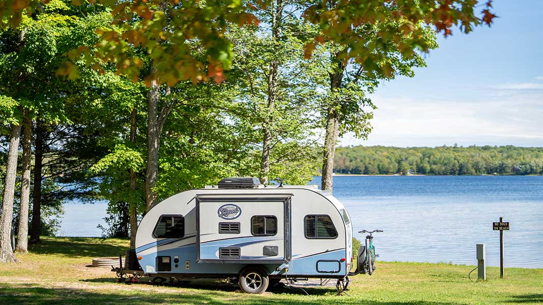 An RV camper sites by a lake at a park with fall color on the trees