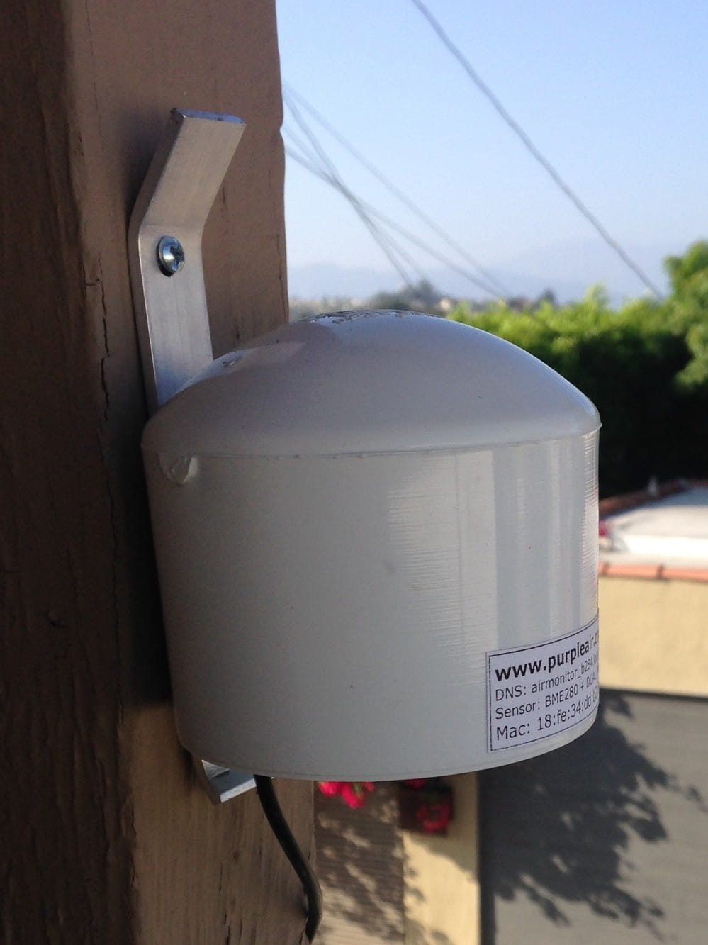 A small white metal cannister (a PurpleAir Monitor) nailed to a wall.
