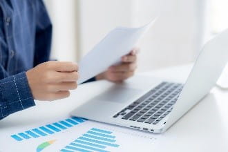 Person holding paper at desk hovering over laptop