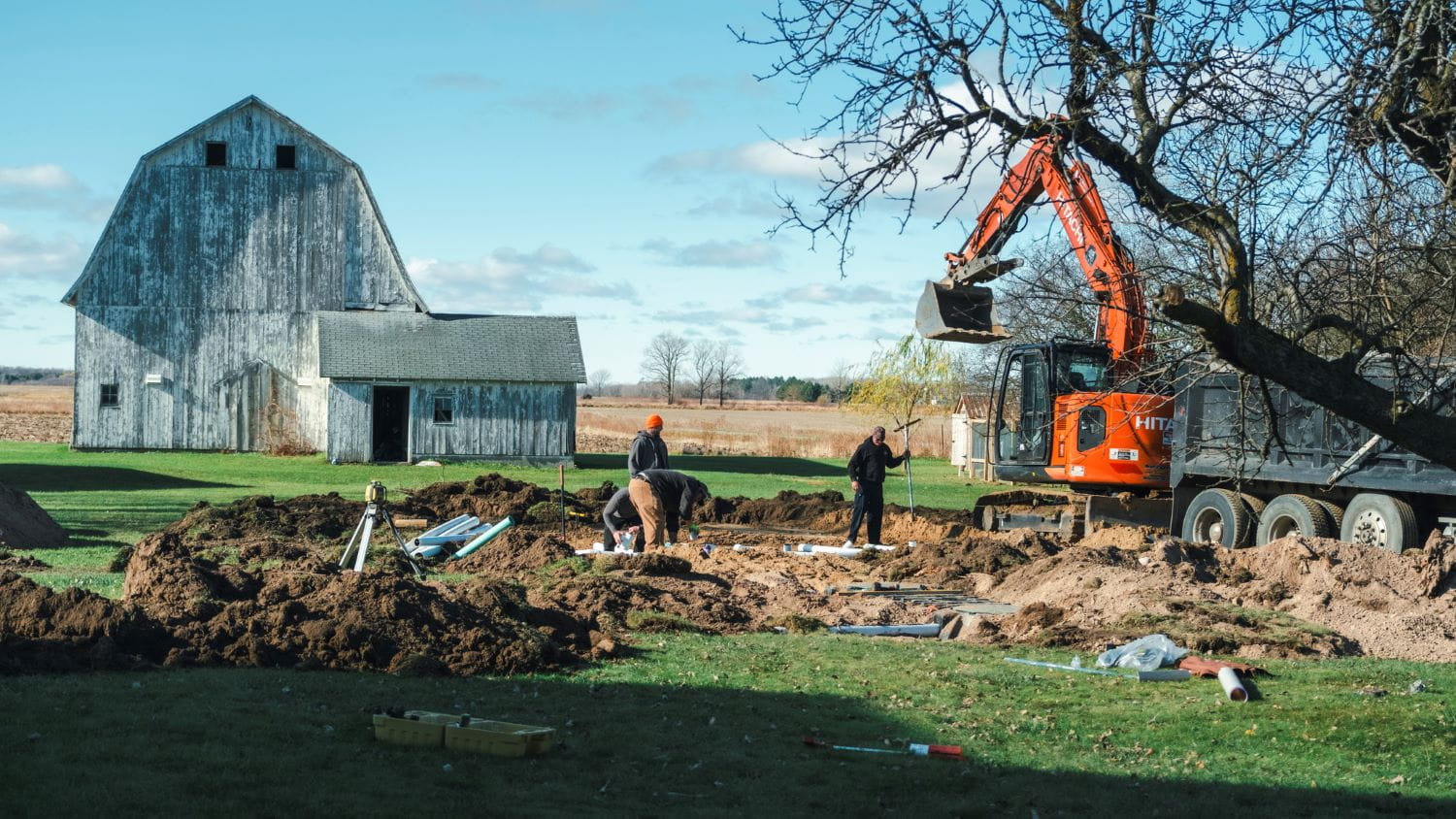 Large machinery digging into a homeowners yard to replace their septic system with a barn and farm fields in the background