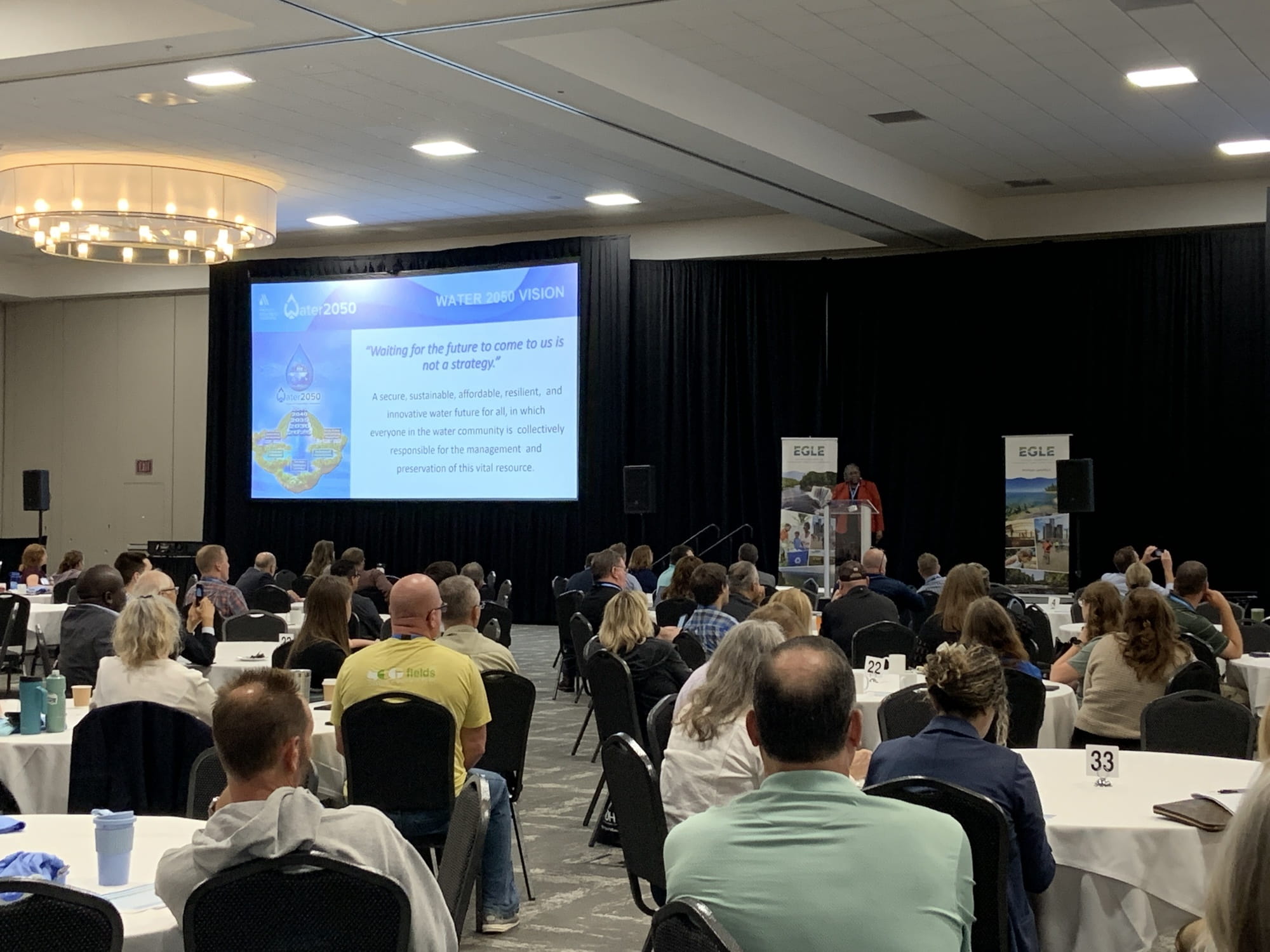 Attendees sitting at tables during a conference watching a presenter on stage