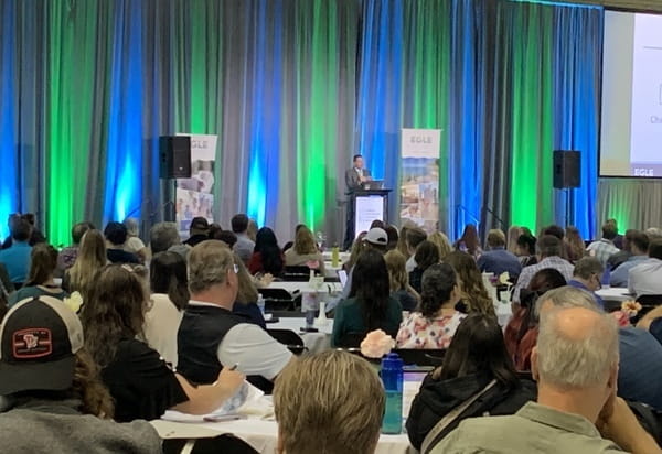 several people sitting at round tables looking at and listening to a speaker on a stage at the 2024 Michigan Environmental Compliance Conference