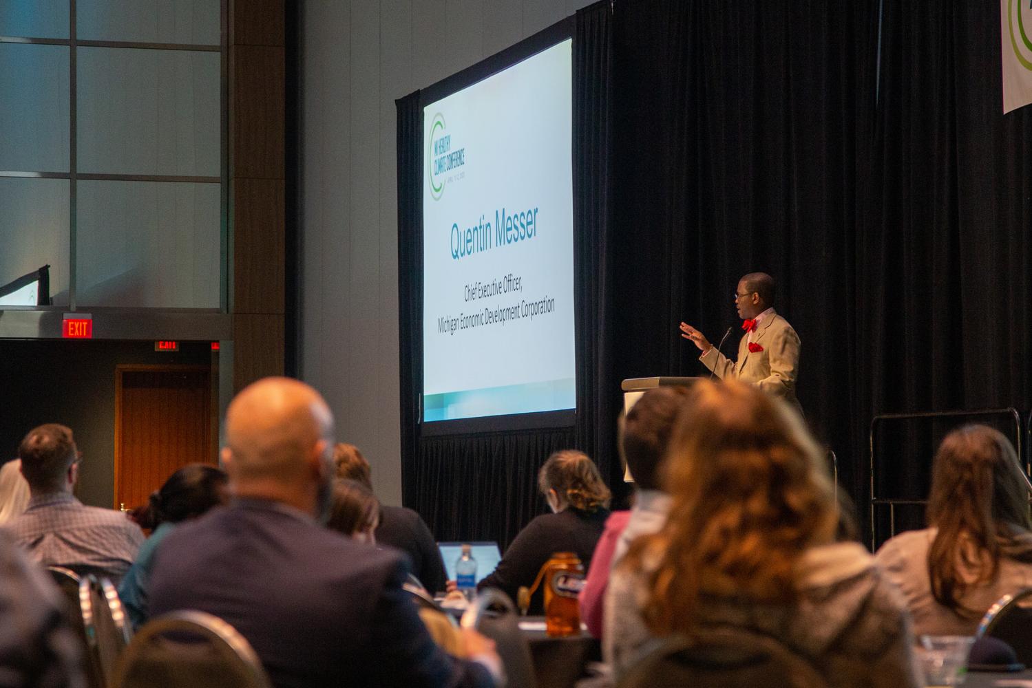A speaker stands at a podium next to a presentation, speaking to a room full of conference attendees
