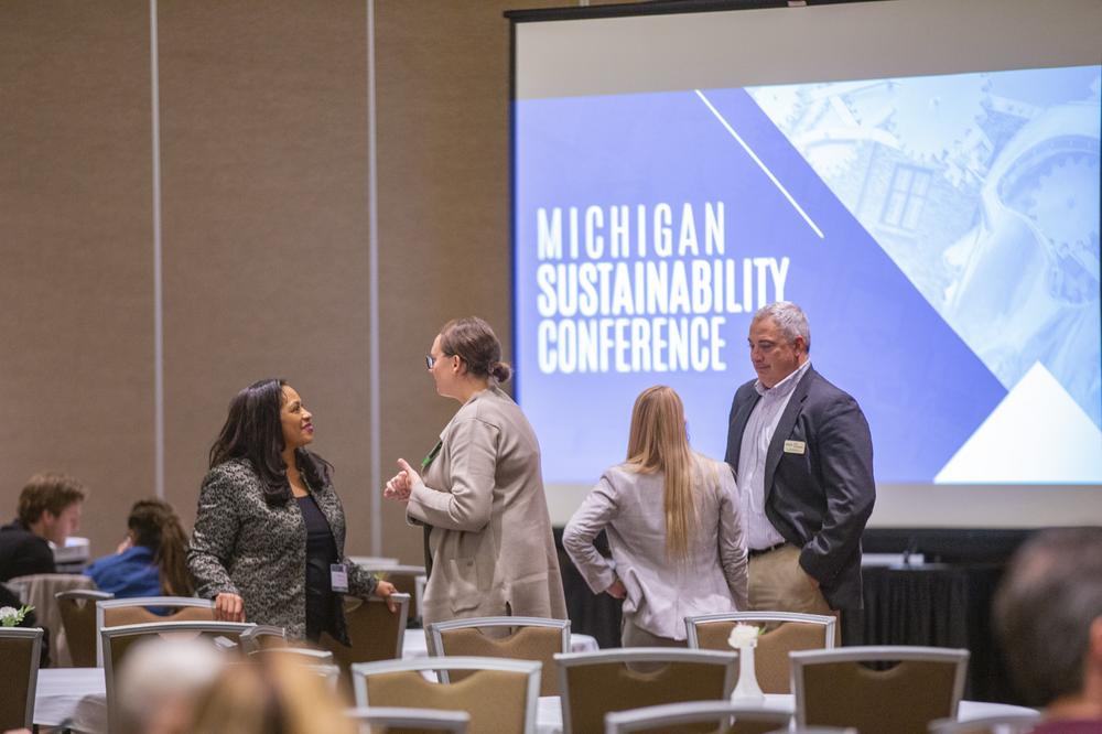 Four people standing in a large presentation room with a projector screen behind them reading "Michigan Sustainability Conference"
