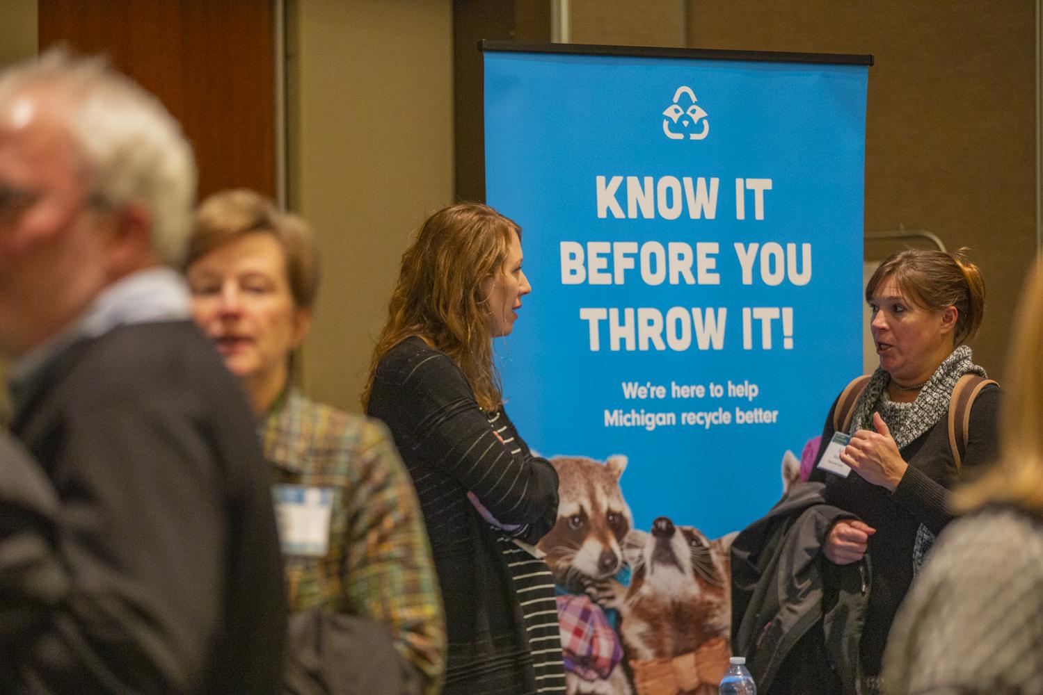 Two female attendees talking in front of a sign that says "Know it before you throw it!"