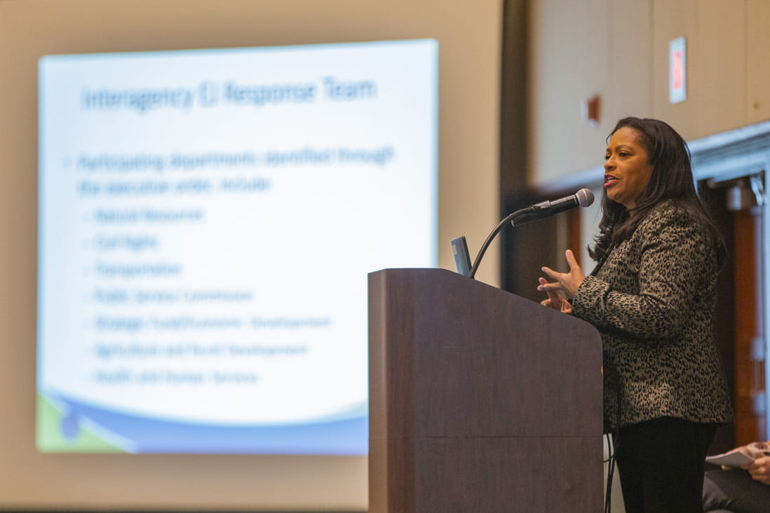 Regina Strong at a podium, gesturing with her hands, with a large presentation slideshow behind her