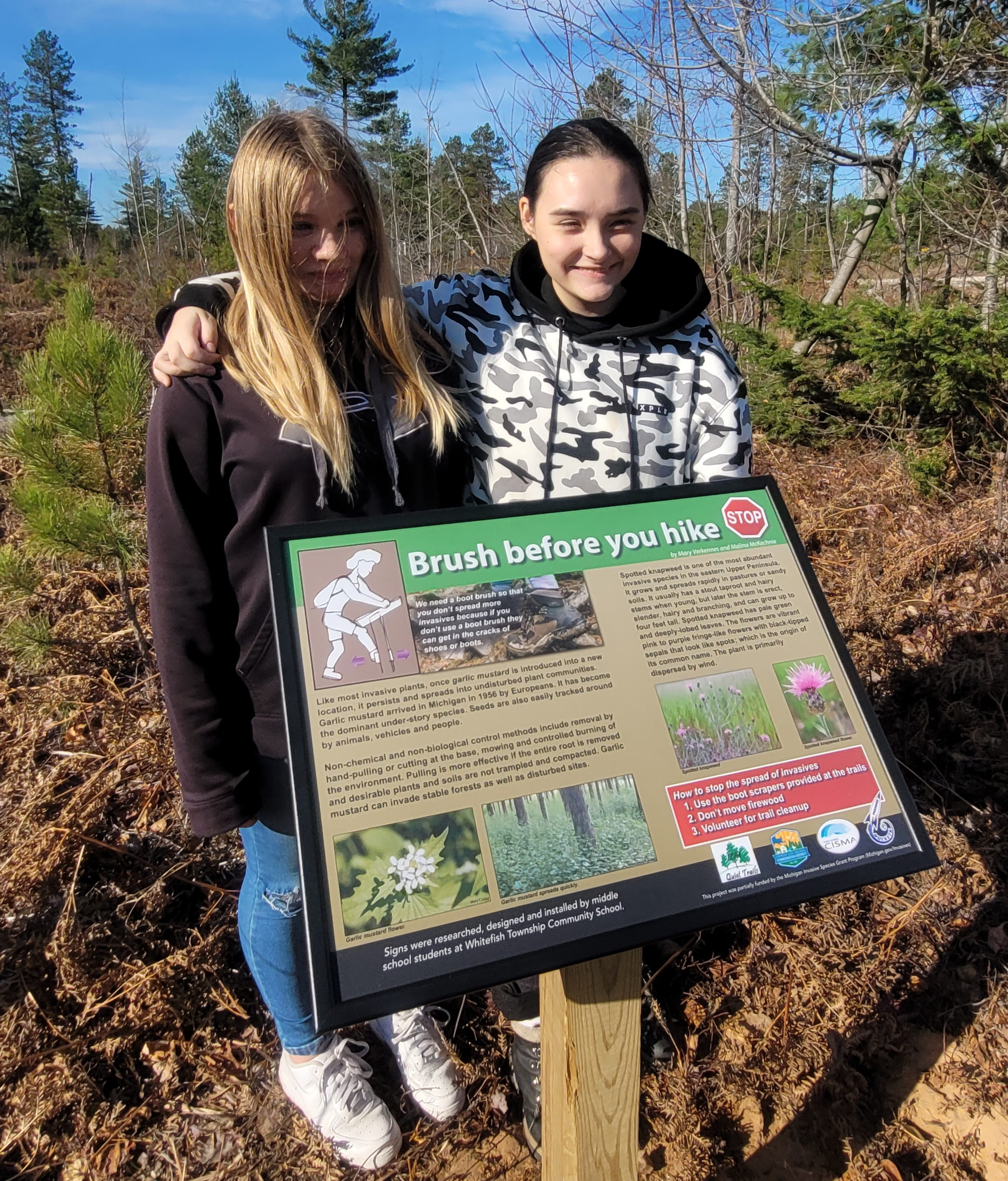 Malina McKechnie, left, and Mary Verkennes, students at Whitefish Township Community School, stand behind "Brush Before You Hike" sign.