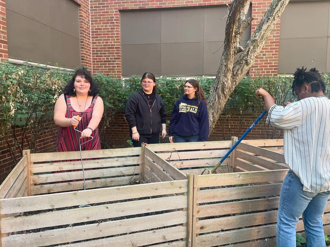 Four high school students stand around wooden compost bins; two students are watching the other two churn the compost piles