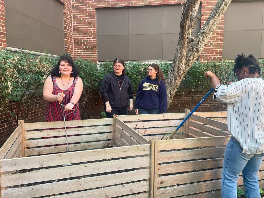 Four high school students stand around wooden compost bins; two students are watching the other two churn the compost piles