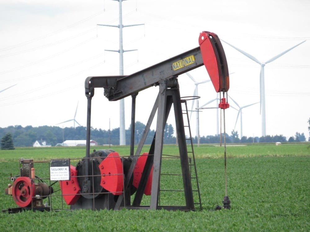 A red oil drilling pump in a grass field with windmills in the background