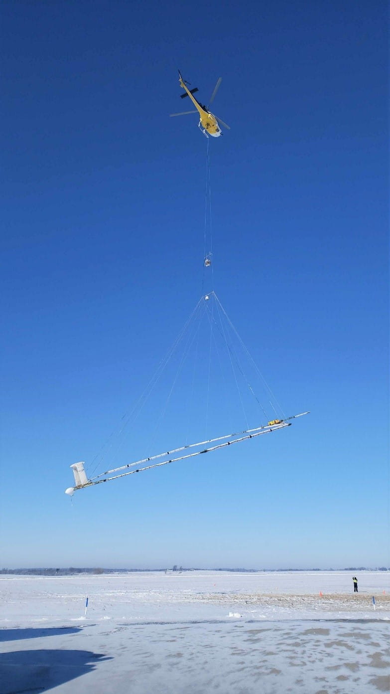 Helicopter with a geophysical equipment loop deployed below it via slingload used for airborne electromagnetic (AEM) surveys. Technician on ground for scale.