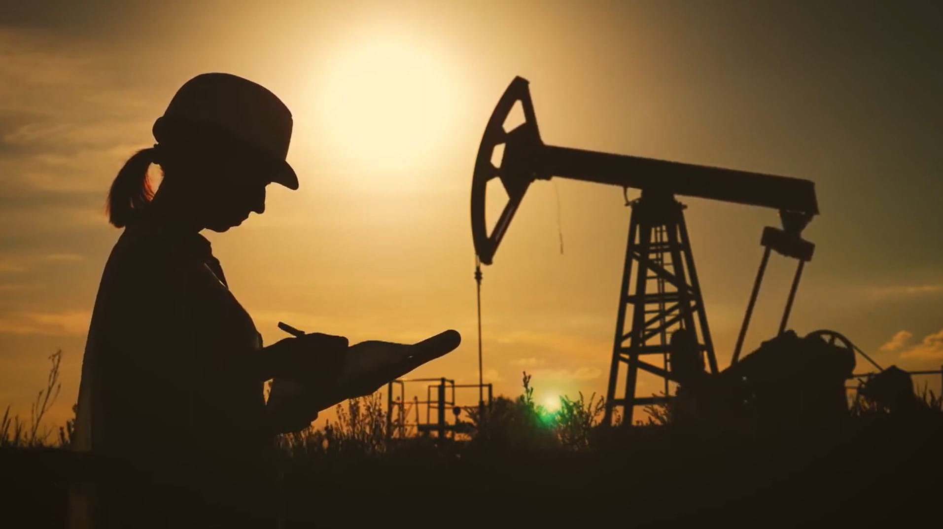 sunsetting behind a person in a hard hat with a clipboard next to an oil rig