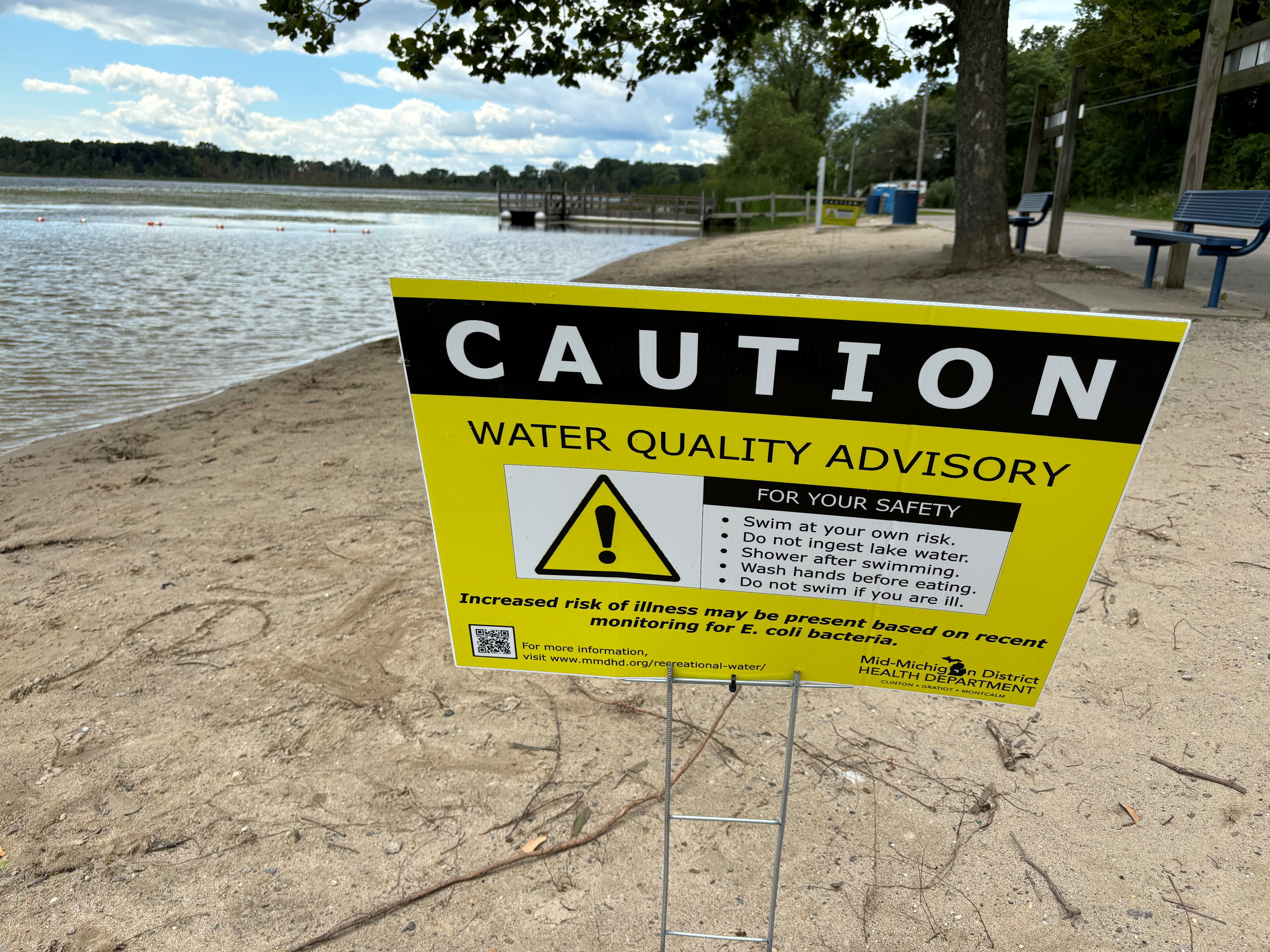 Water quality advisory sign on a sandy lakeshore, cautioning against swimming due to increased risk of illness due to E. coli