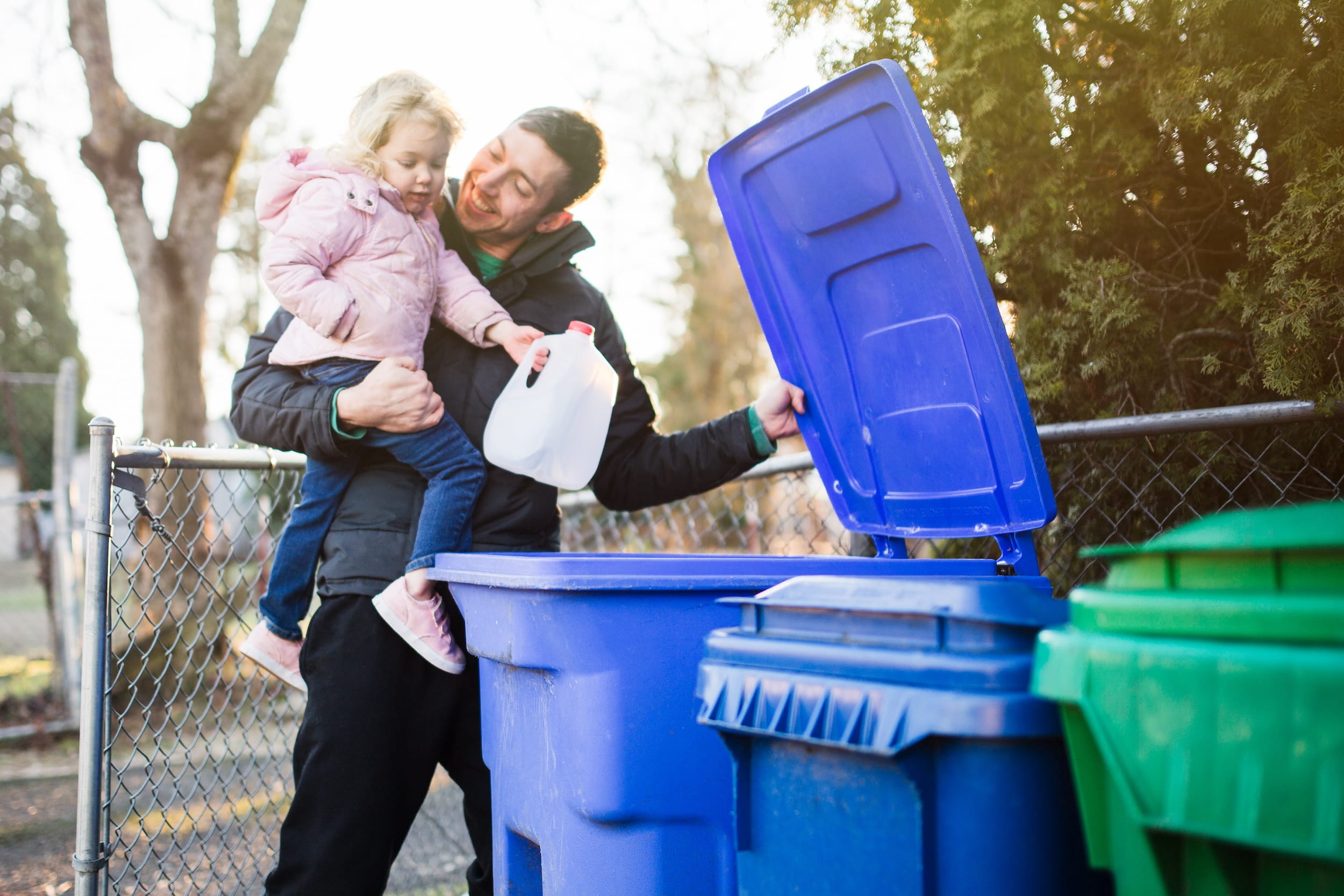 Girl held by her father places milk carton into recycling cart. 