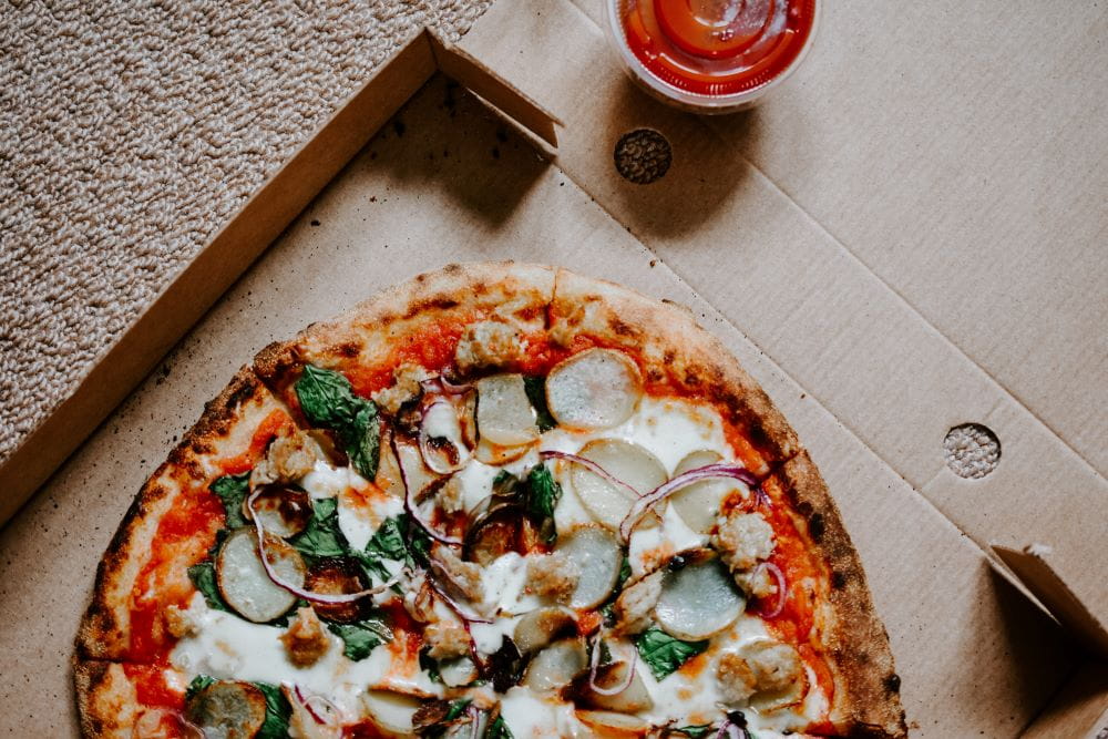 A pizza sitting in a greasy pizza box, a cup of marinara sauce nearby