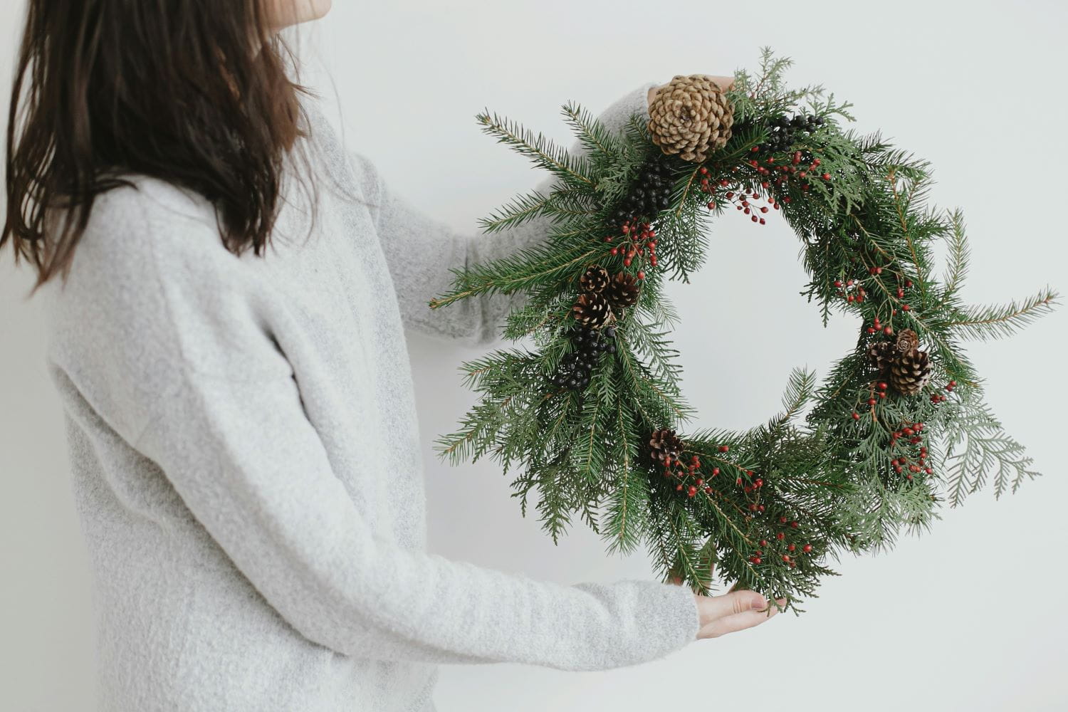 A person holding an evergreen wreath decorated with berries and pinecones