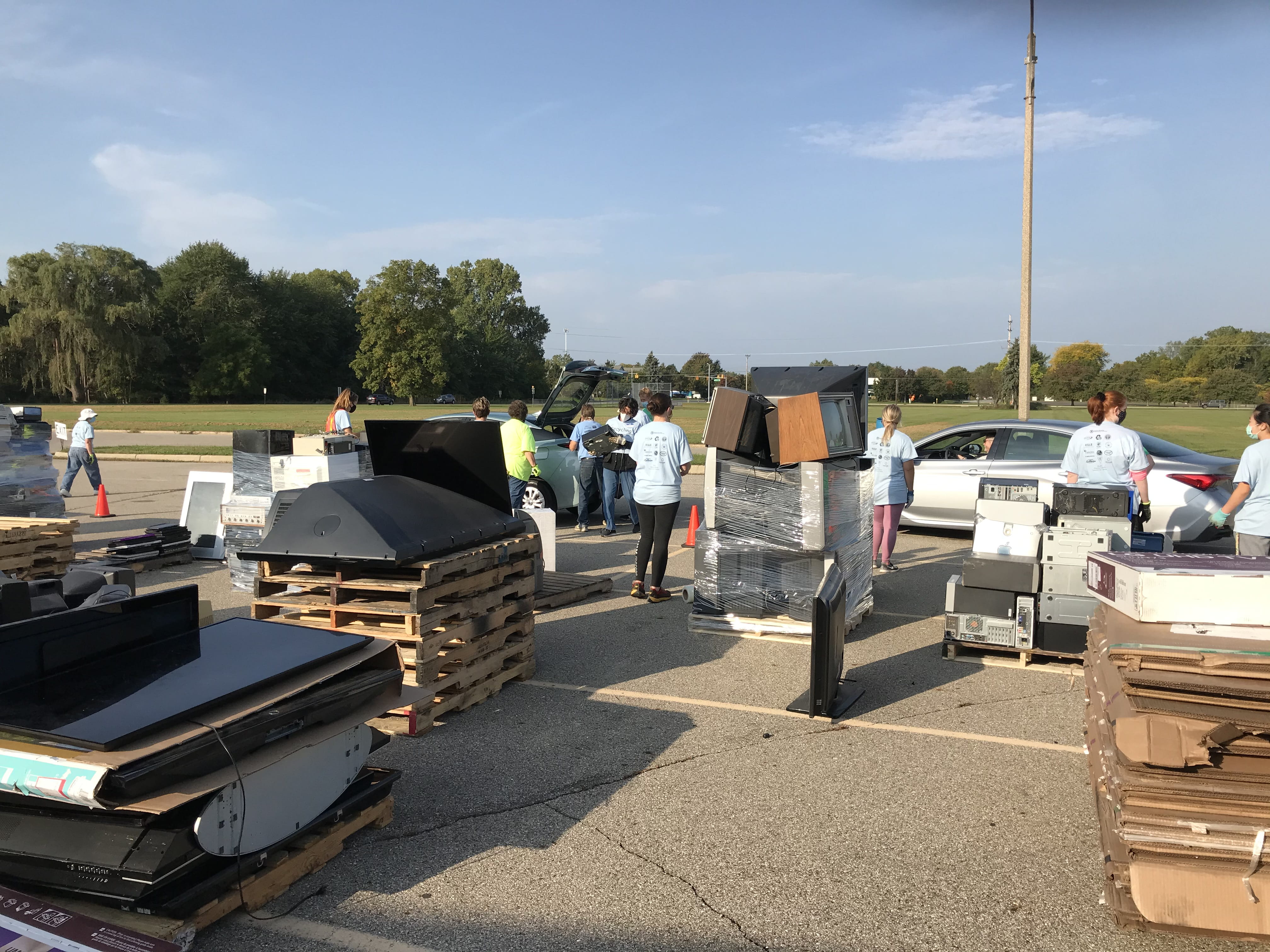 Community members at a drive-thru electronic recycling event. Volunteers are assisting residents pulling up in their cars in a parking lot.