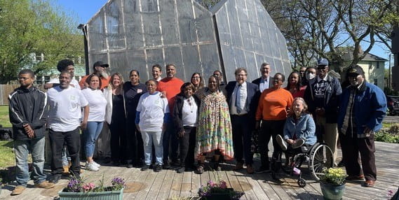 A diverse group of state and local leaders gathered in front of an abstract art installation, smiling on a sunny spring day