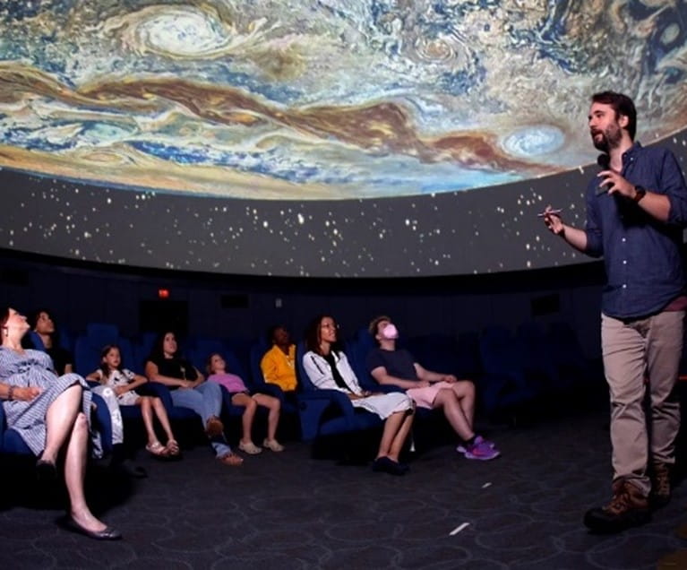 Visitors sitting down for a tour at the Longway Planetarium in a dark room with a planet on the ceiling.