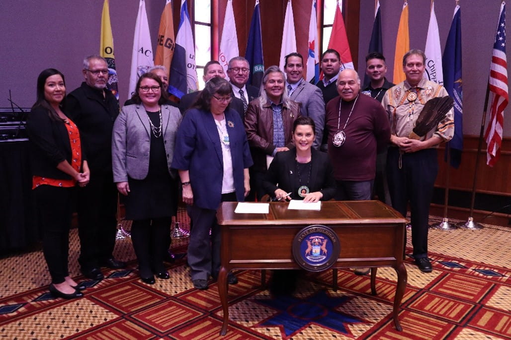 Governor Gretchen Whitmer sits at a desk in front of Tribal leaders, signing Executive Directive 2019-17