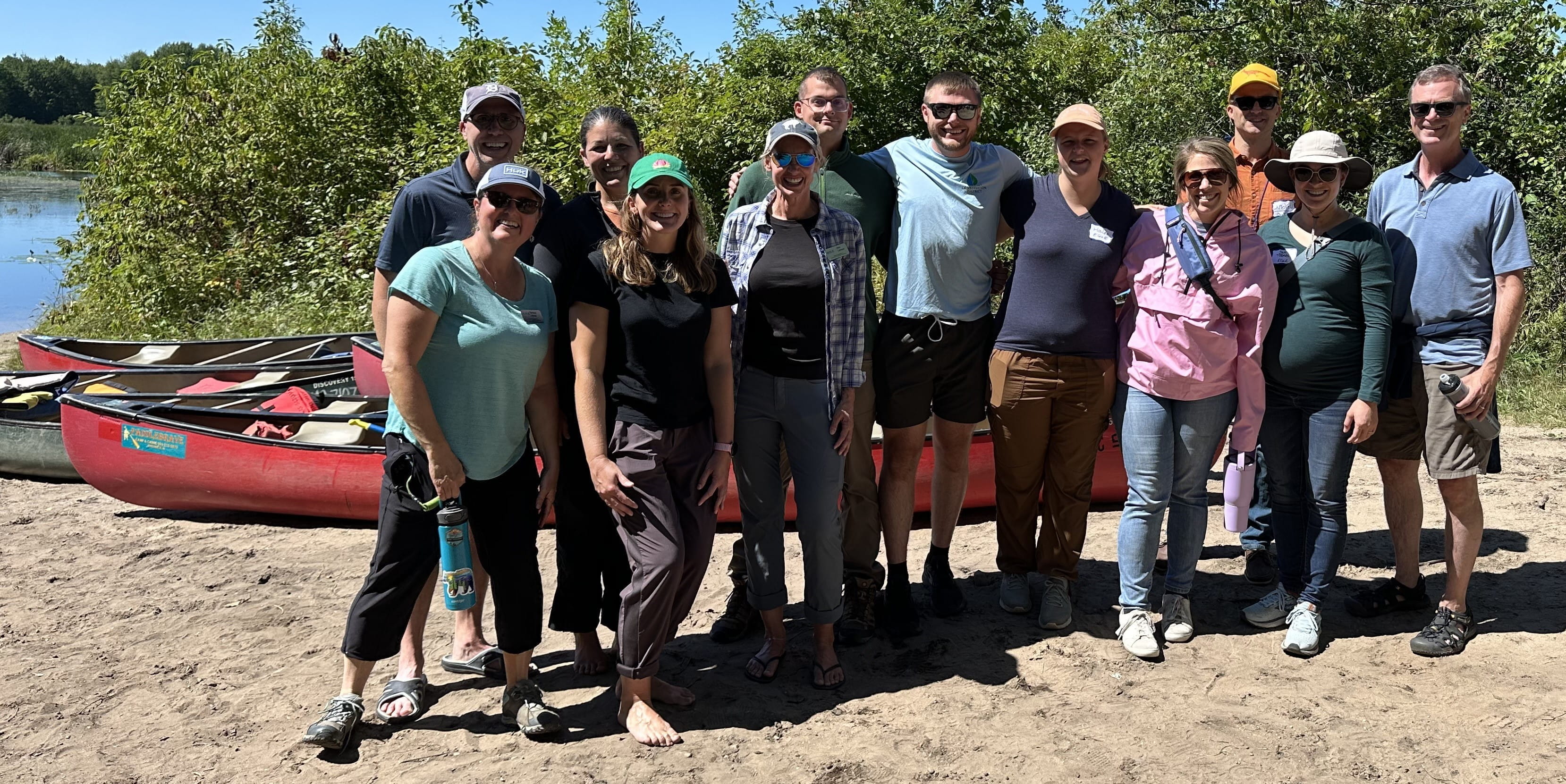 A group of 12 EGLE staff, huddled together in front of canoes, smiling in the summer sunshine